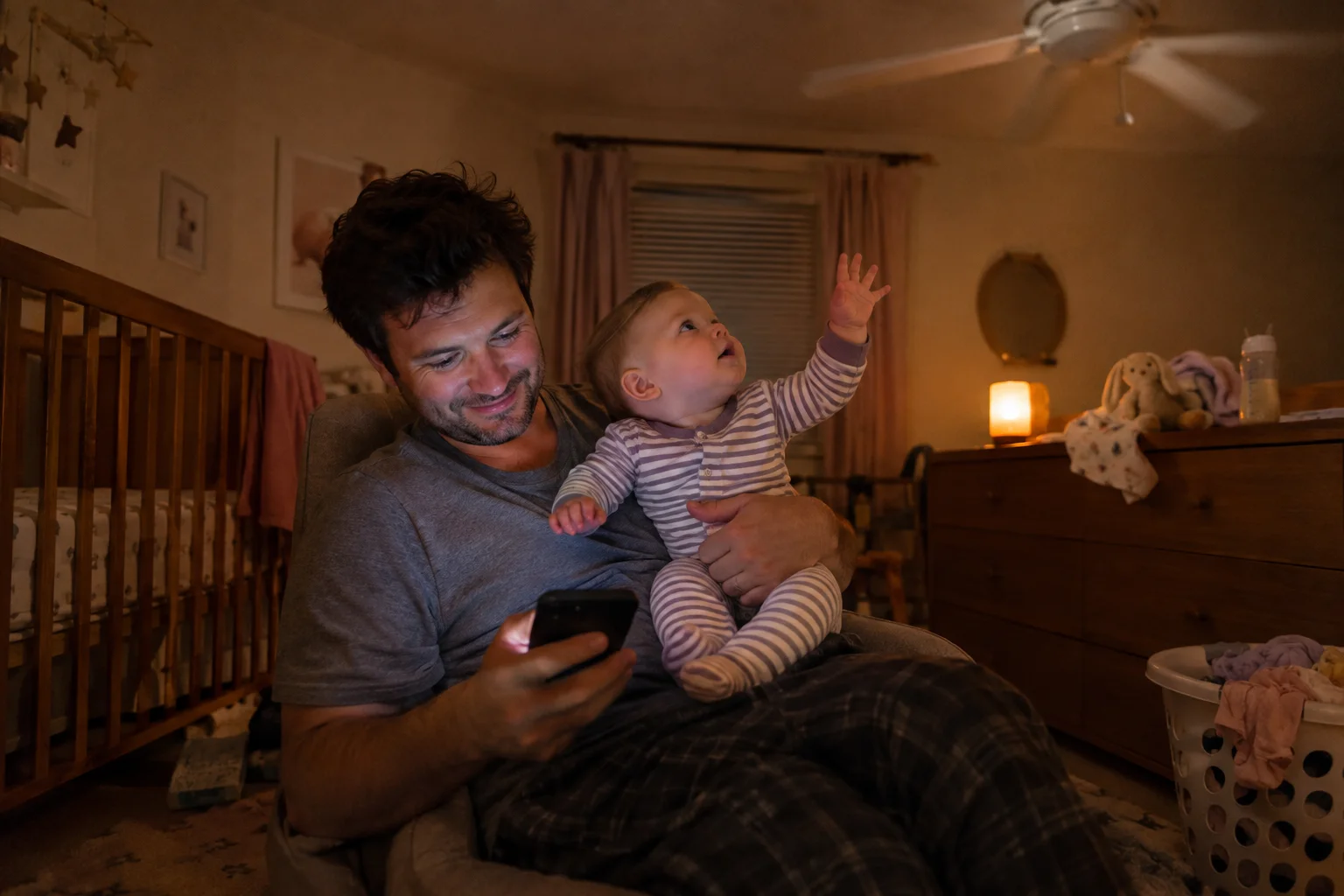 A dad holds a phone and a baby who reaches toward a ceiling fan in a softly lit nursery.