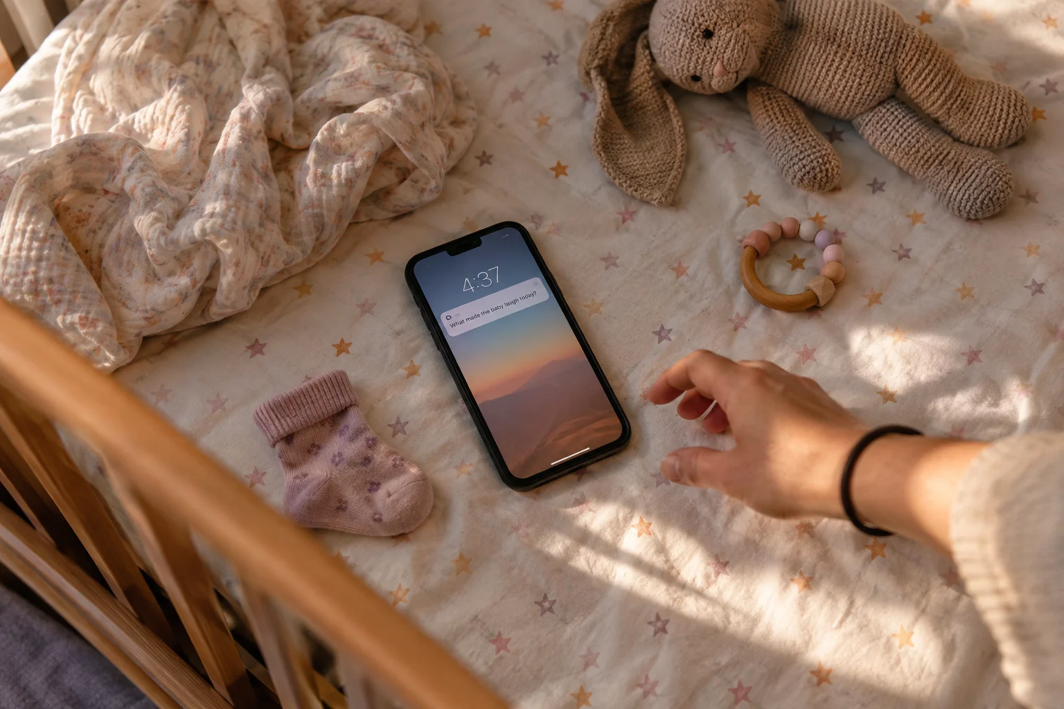A phone with a notification lies on a patterned crib sheet beside a plush bunny and a reaching hand.